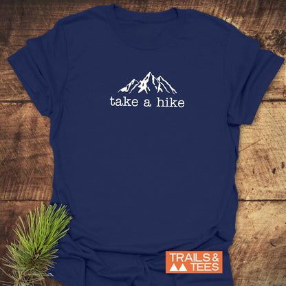 Take A Hike T-Shirt featuring a white mountain graphic and take a hike text on navy-blue fabric, styled with a pine twig, showcasing its outdoor-inspired design and comfort.