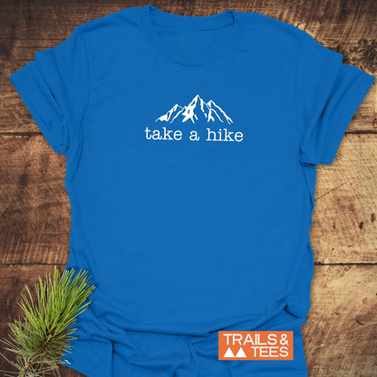 Take A Hike T-Shirt featuring a mountain range line-drawing and take a hike text on chest, laid flat with a small pine sprig and an orange TRAILS & TEES logo.
