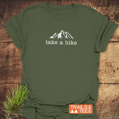 Take A Hike T-Shirt featuring a mountain outline and slogan, set on a wooden background with pine sprig, emphasizing outdoor adventure. Displayed with Trails & Tees logo, highlighting comfort and durability.