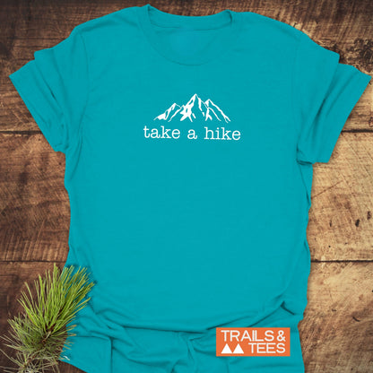 Take A Hike T-Shirt featuring a white mountain line-drawing and take a hike text, displayed on a wooden surface with a small orange TRAILS & TEES badge and pine needles nearby.