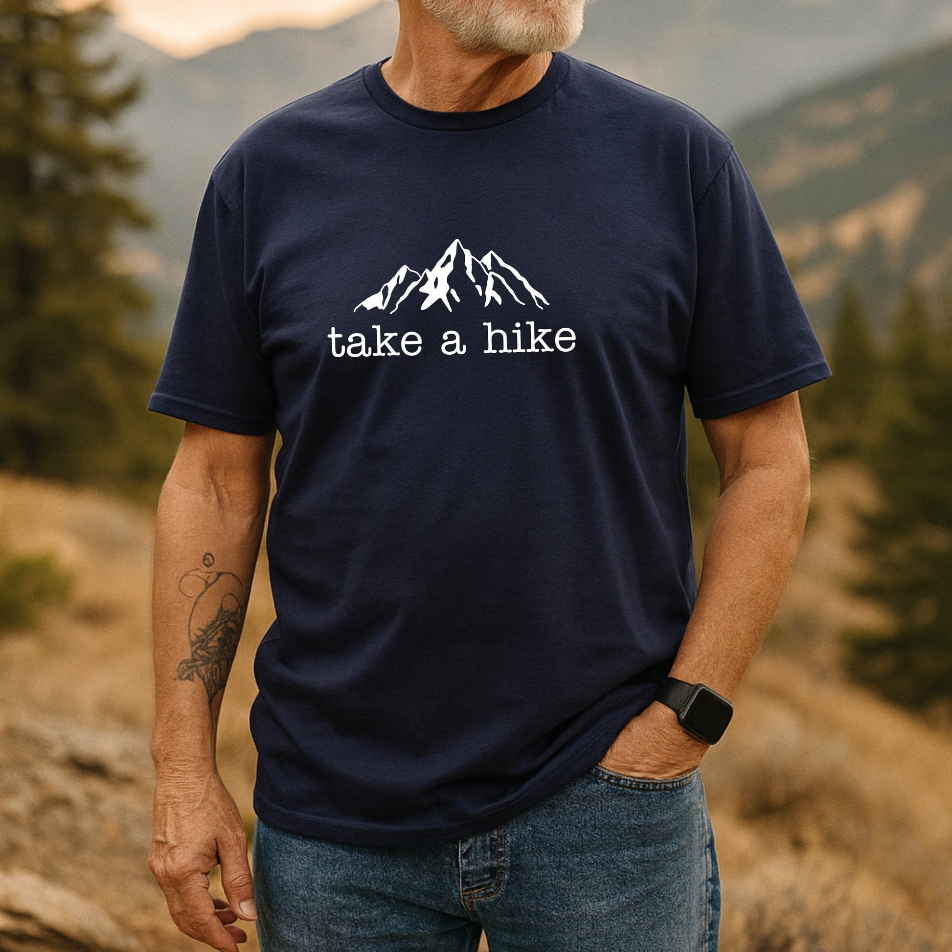 Person wearing the Take A Hike T-Shirt, featuring a mountain silhouette and slogan. The dark T-shirt showcases durable prints, perfect for outdoor enthusiasts, complemented by a smartwatch and visible tattoo.