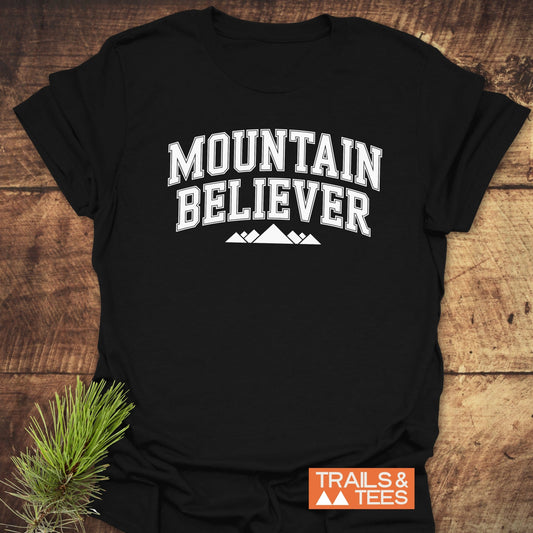 Mountain Believer T-Shirt on wooden surface with bold white collegiate text and mountain graphic. Pine sprig decorates lower left; orange Trails & Tees logo at bottom right.