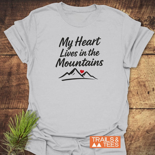 My Heart Lives In The Mountains T-Shirt featuring stylized mountain peaks and a red heart graphic, laid flat on a wood surface with pine sprig, capturing an outdoorsy, nature-loving theme.