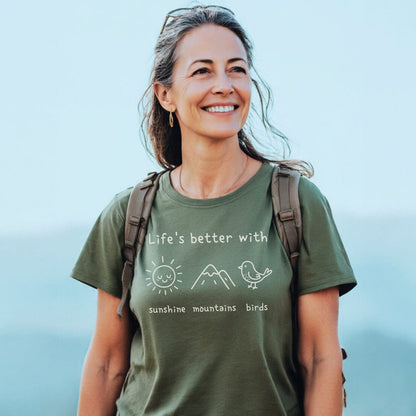 A woman wears the Sunshine Mountains Birds T-Shirt, featuring a slogan and nature icons, while outdoors. Her green tee highlights a love for the outdoors, embodying comfort and adventure.