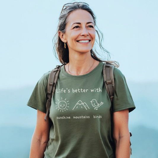 A woman wears the Sunshine Mountains Birds T-Shirt, featuring a slogan and nature icons, while outdoors. Her green tee highlights a love for the outdoors, embodying comfort and adventure.