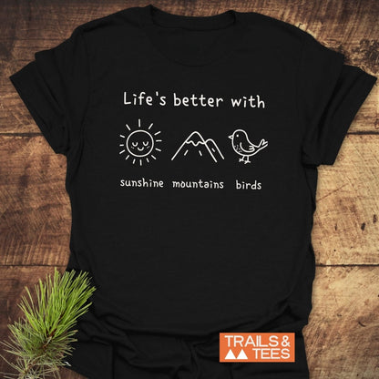 Sunshine Mountains Birds T-Shirt with white print of sun, mountains, and bird icons, plus TRAILS & TEES logo on a wooden background, emphasizing outdoor adventure and comfort.