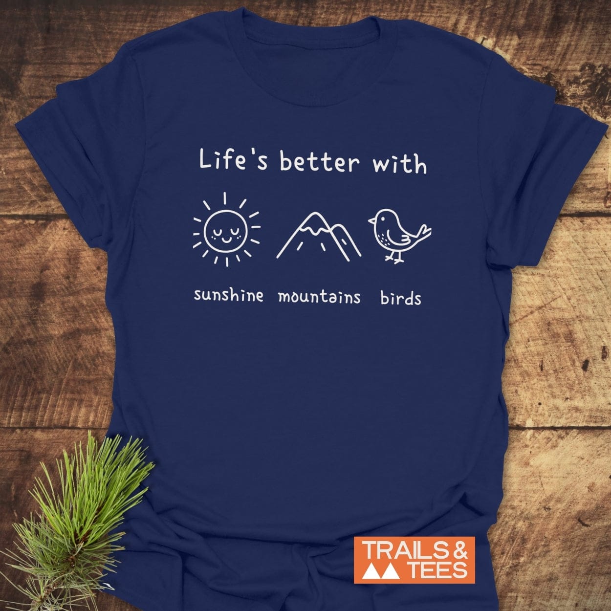 Sunshine Mountains Birds T-Shirt on a wooden surface, featuring a white line-drawing with sun, mountain, and bird icons. Includes TRAILS & TEES logo, reflecting outdoor adventure spirit.