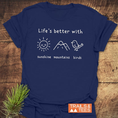 Sunshine Mountains Birds T-Shirt on a wooden surface, featuring a white line-drawing with sun, mountain, and bird icons. Includes TRAILS & TEES logo, reflecting outdoor adventure spirit.