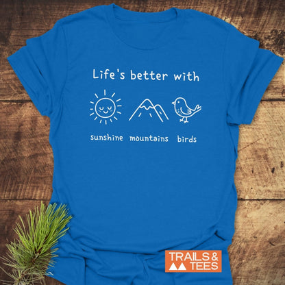 Sunshine Mountains Birds T-Shirt featuring hand-drawn icons of a sun, mountain, and bird with labels, laid flat on a wooden surface. Includes TRAILS & TEES logo in the corner.