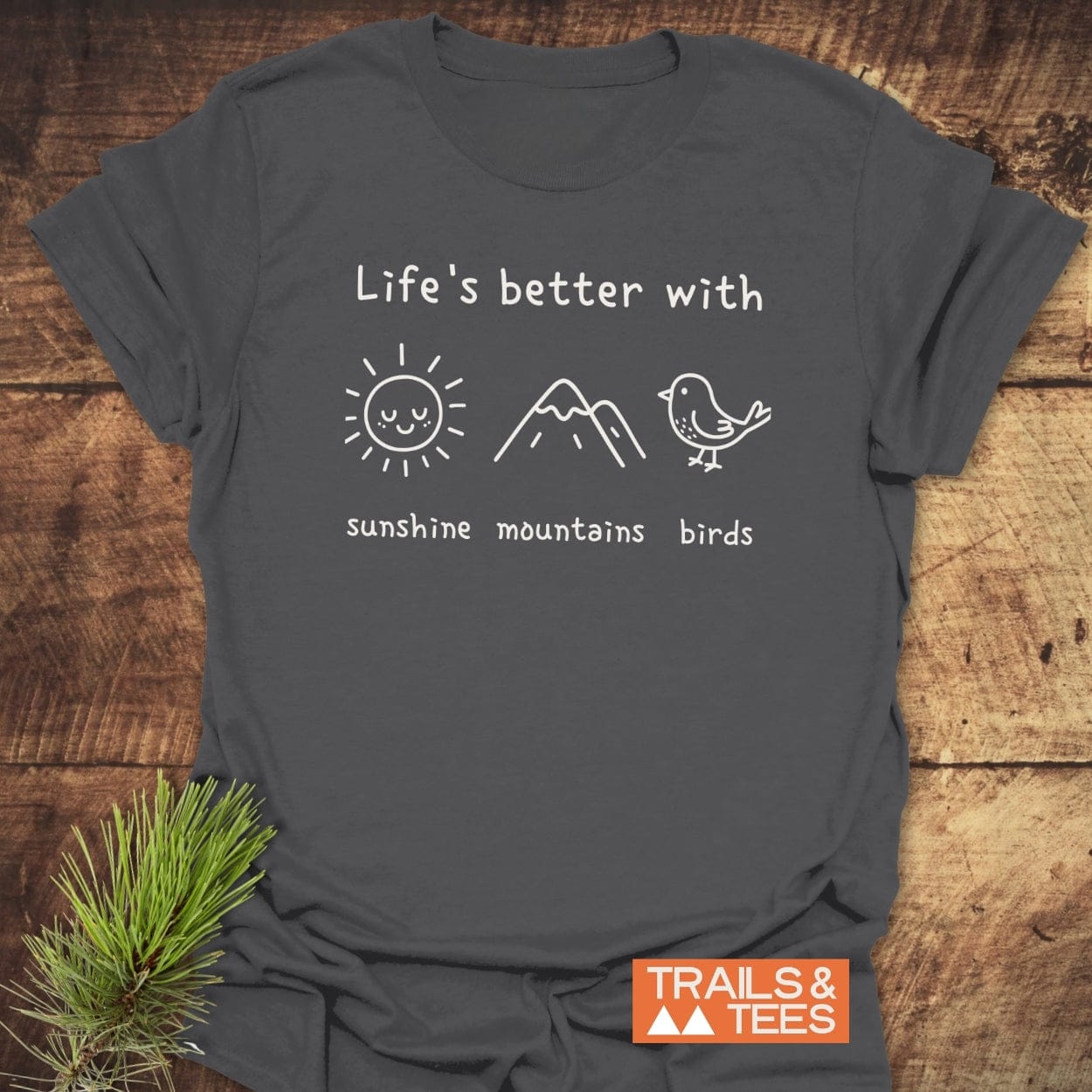 Sunshine Mountains Birds T-Shirt in charcoal-gray, featuring white hand-drawn doodles of a sun, mountains, and birds. Laid on wood with a pine sprig, showcasing outdoor adventure spirit.