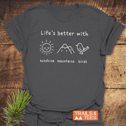 Sunshine Mountains Birds T-Shirt in charcoal-gray, featuring white hand-drawn doodles of a sun, mountains, and birds. Laid on wood with a pine sprig, showcasing outdoor adventure spirit.