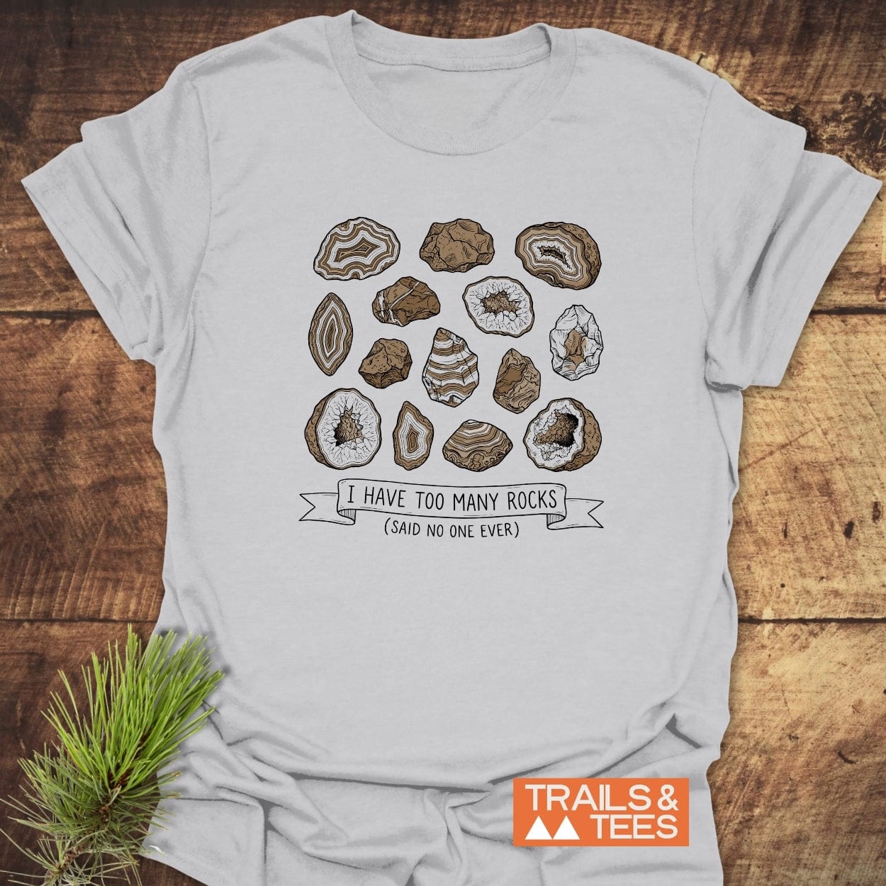 I Have Too Many Rocks T-Shirt featuring stylized geode slices and a witty slogan, ideal for outdoor enthusiasts. Crafted from soft ring-spun cotton with durable prints, showcasing the Trails & Tees logo.