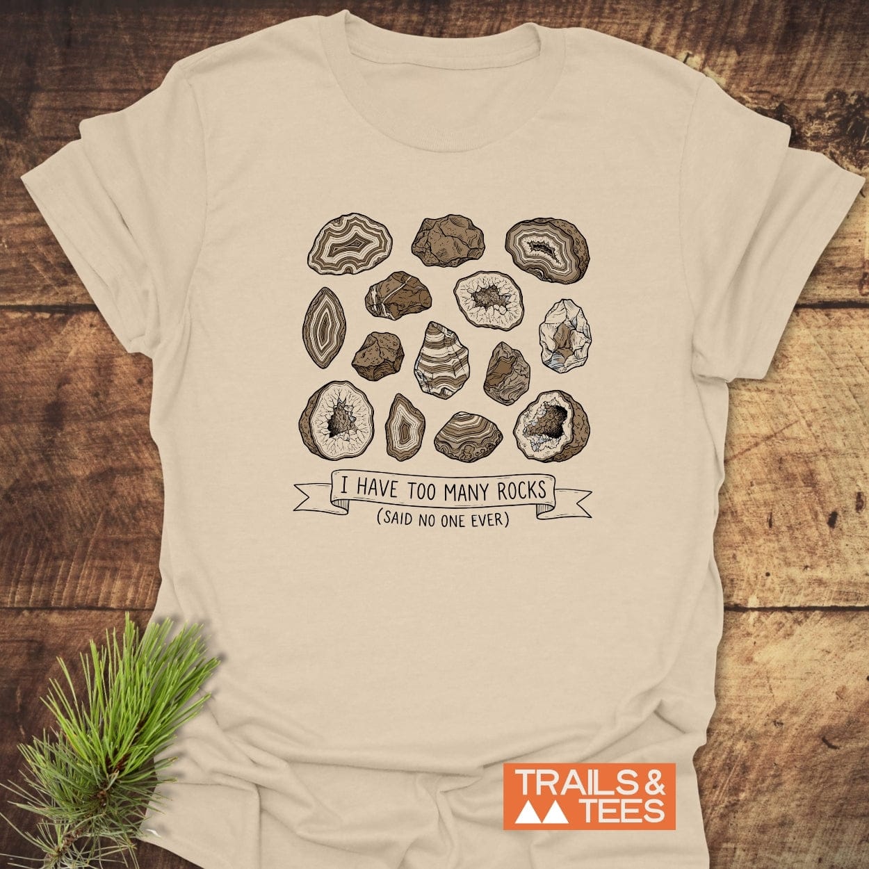 I Have Too Many Rocks T-Shirt featuring hand-drawn rock illustrations and playful slogan, laid on wood. Crafted from soft, lightweight cotton, showcasing Trails & Tees logo with pine sprig detail.