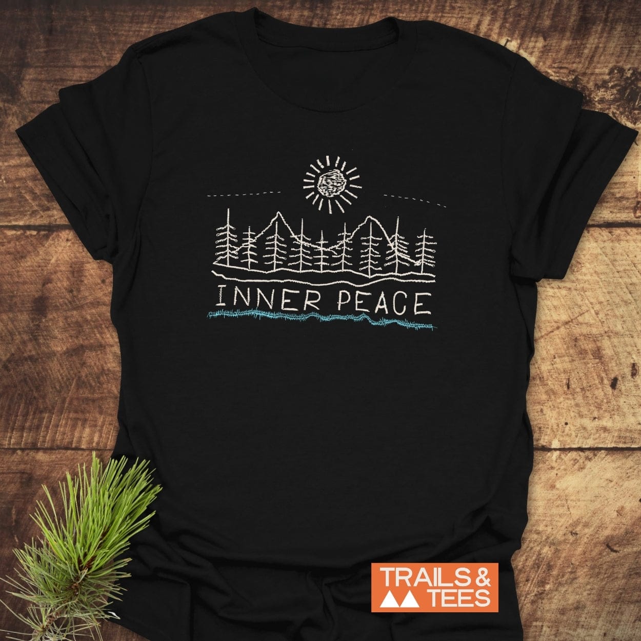Inner Peace T-Shirt features a hand-drawn scene of pine trees and mountains beneath a sun, with 'INNER PEACE' text. Includes 'TRAILS & TEES' logo and pine sprig, emphasizing outdoor adventure.