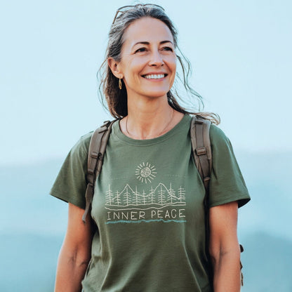 Smiling woman outdoors wearing the Inner Peace T-Shirt, featuring tree and sun line-drawing. Green tee, backpacked, with sunglasses on her head, small hoop earrings, embodying outdoor adventure spirit.