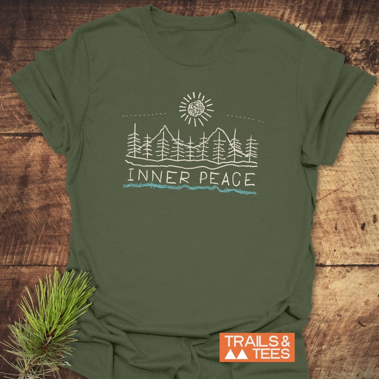 Inner Peace T-Shirt: Olive-green short-sleeve tee with a sun, pine trees, and mountains graphic, “INNER PEACE” text below. Styled with a pine sprig, highlighting outdoor adventure spirit.