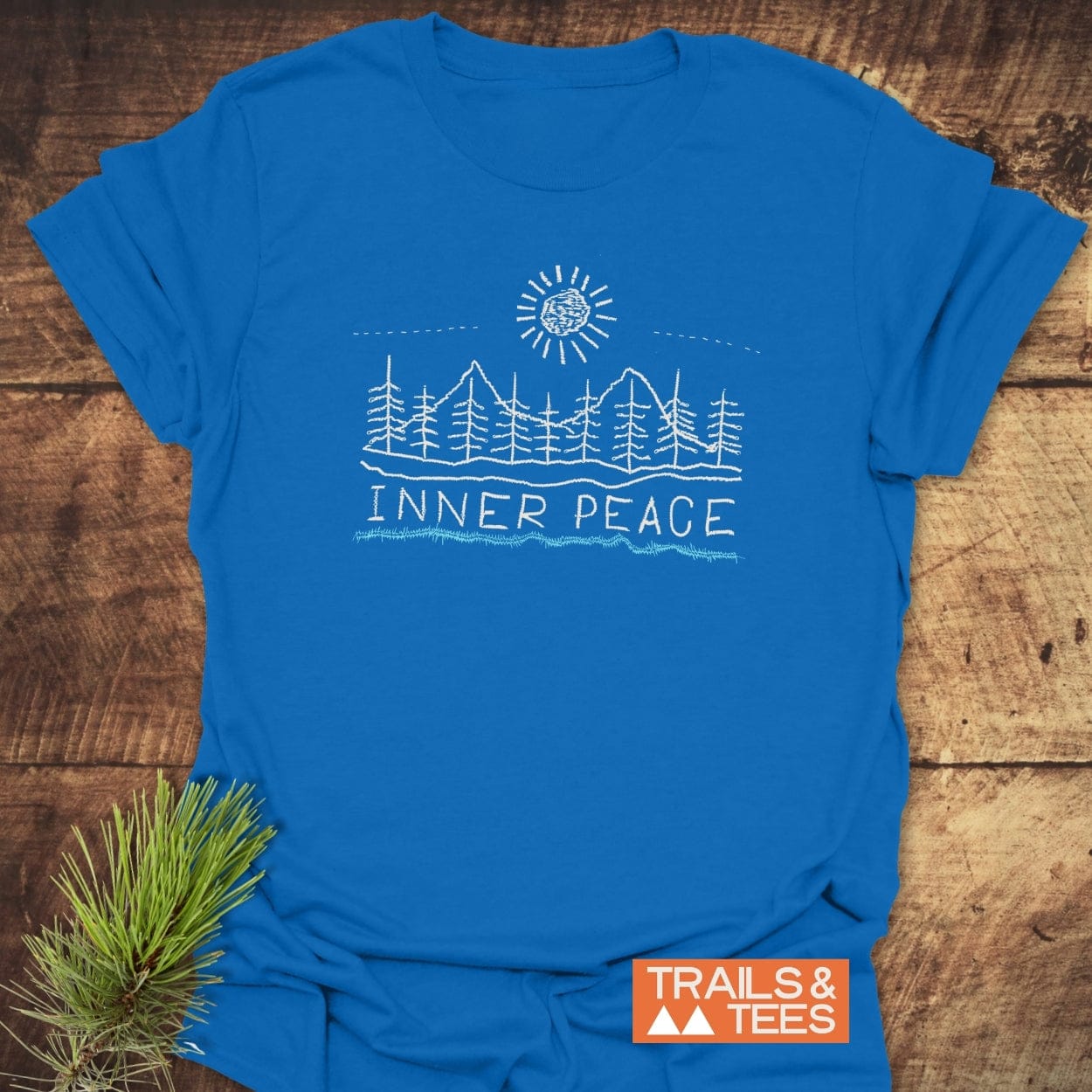 Inner Peace T-Shirt featuring a line-art scene of pine trees and mountains under a stylized sun. Laid flat with a pine sprig beside it, showcasing Trails & Tees logo for an outdoor vibe.