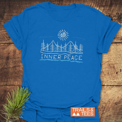 Inner Peace T-Shirt featuring a line-art scene of pine trees and mountains under a stylized sun. Laid flat with a pine sprig beside it, showcasing Trails & Tees logo for an outdoor vibe.