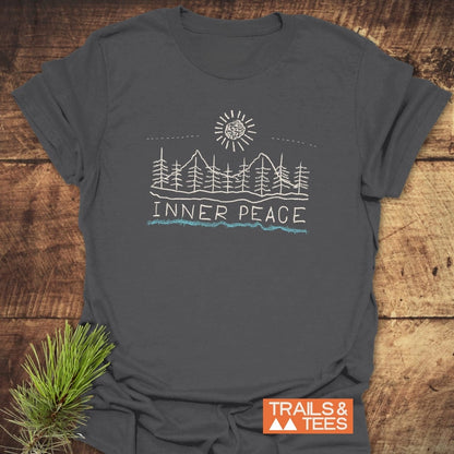 Inner Peace T-Shirt featuring hand-drawn mountains, pine trees, and sun graphic with INNER PEACE text, light blue underline, and orange TRAILS & TEES logo, on charcoal-gray cotton.