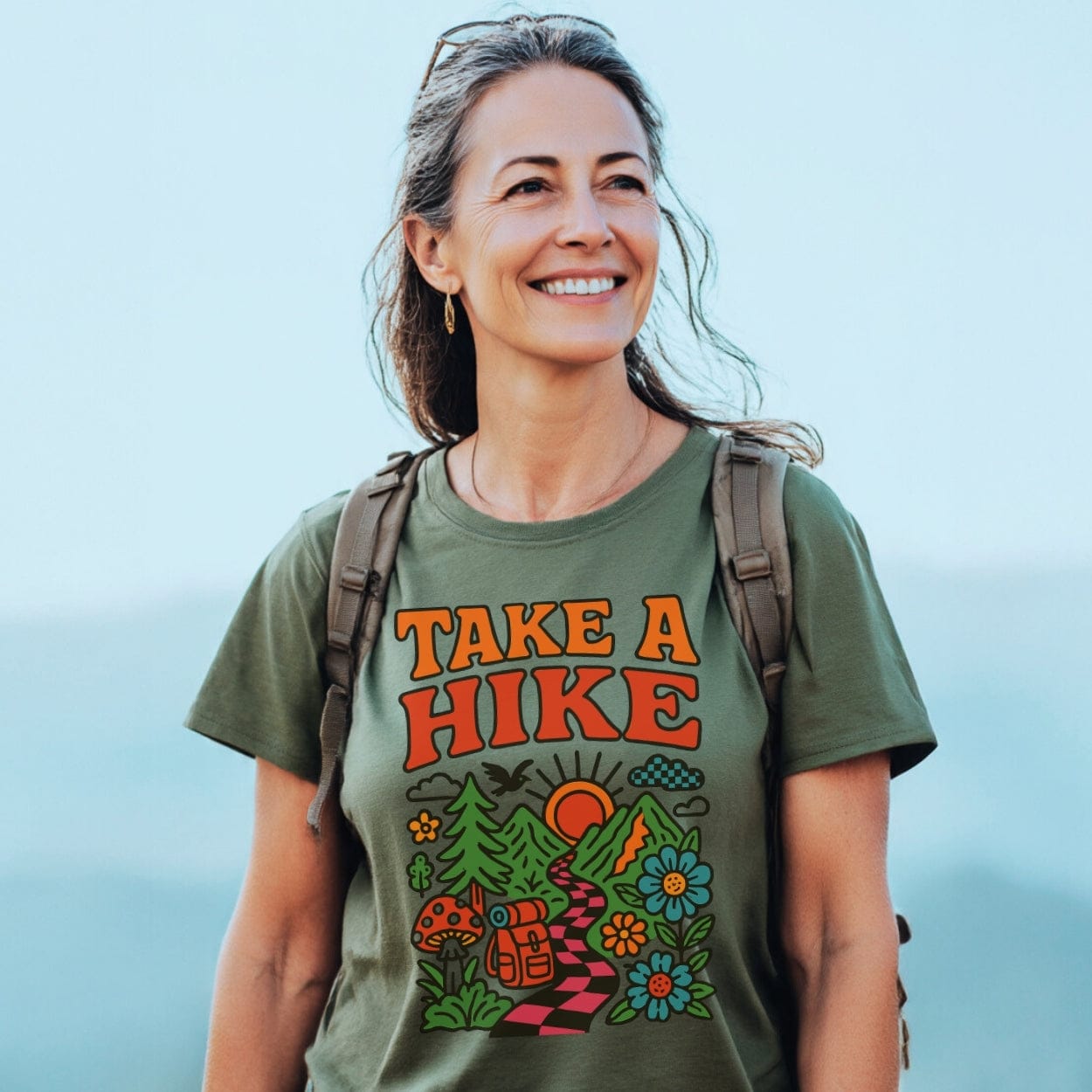 Take A Hike Funky T-Shirt featuring a retro TAKE A HIKE design with mountains and trees, worn by a smiling woman outdoors, highlighting a love for hiking and nature.
