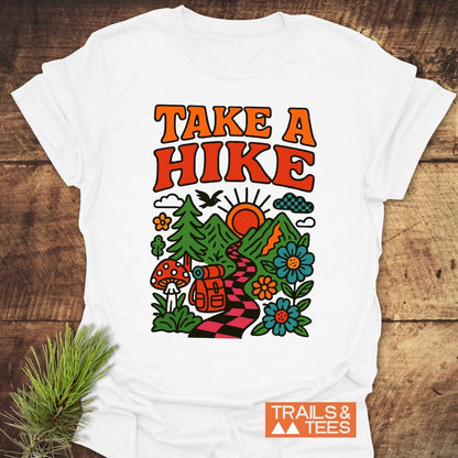 Take A Hike Funky T-Shirt features retro “TAKE A HIKE” text above a vibrant hiking scene with mountains, sun, trees, and a trail. Includes “TRAILS & TEES.” logo. Crafted for comfort and adventure.