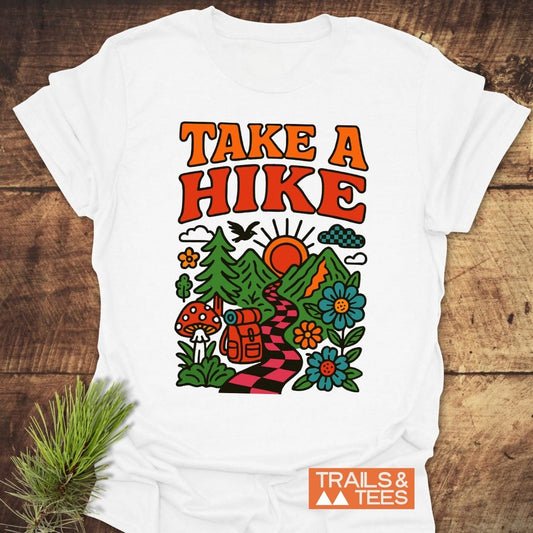 Take A Hike Funky T-Shirt features retro “TAKE A HIKE” text above a vibrant hiking scene with mountains, sun, trees, and a trail. Includes “TRAILS & TEES.” logo. Crafted for comfort and adventure.