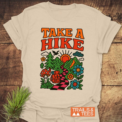 Beige Take A Hike Funky T-Shirt featuring retro design with sun, mountains, trail, backpack, and nature elements. Includes TRAILS & TEES logo on rustic wood background with pine sprig.