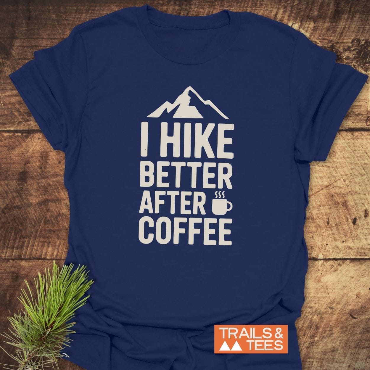 Navy-blue I Hike Better After Coffee T-shirt with mountain and coffee-cup graphic, laid on wood, featuring TRAILS & TEES logo, and accompanied by a pine sprig for an outdoorsy touch.