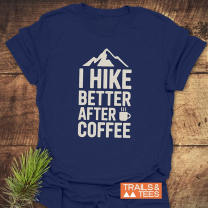 Navy-blue I Hike Better After Coffee T-shirt with mountain and coffee-cup graphic, laid on wood, featuring TRAILS & TEES logo, and accompanied by a pine sprig for an outdoorsy touch.