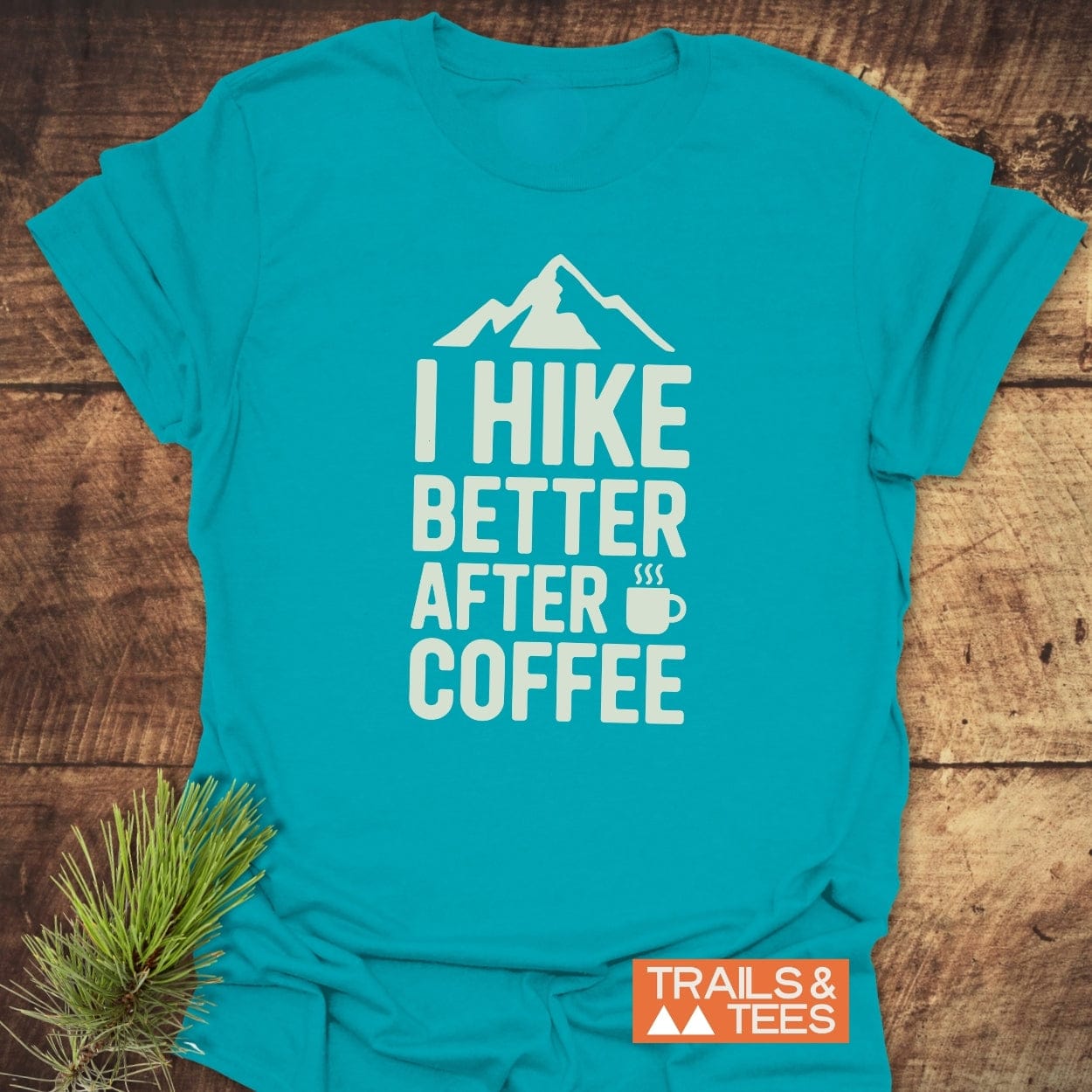 I Hike Better After Coffee T-Shirt featuring a mountain graphic and coffee slogan, showcasing outdoorsy charm. Includes Trails & Tees label, pine sprig accent, highlighting trail-ready design and durability.
