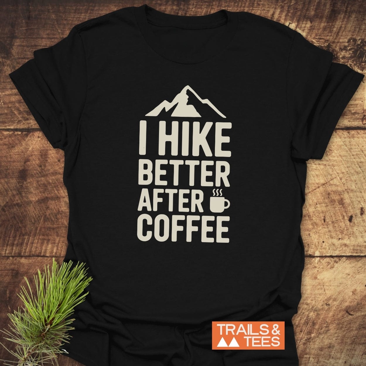 Black T-shirt with I HIKE BETTER AFTER COFFEE text and mountain icon, featuring a steaming coffee cup graphic. Includes TRAILS & TEES logo and pine twig accent on wooden surface.