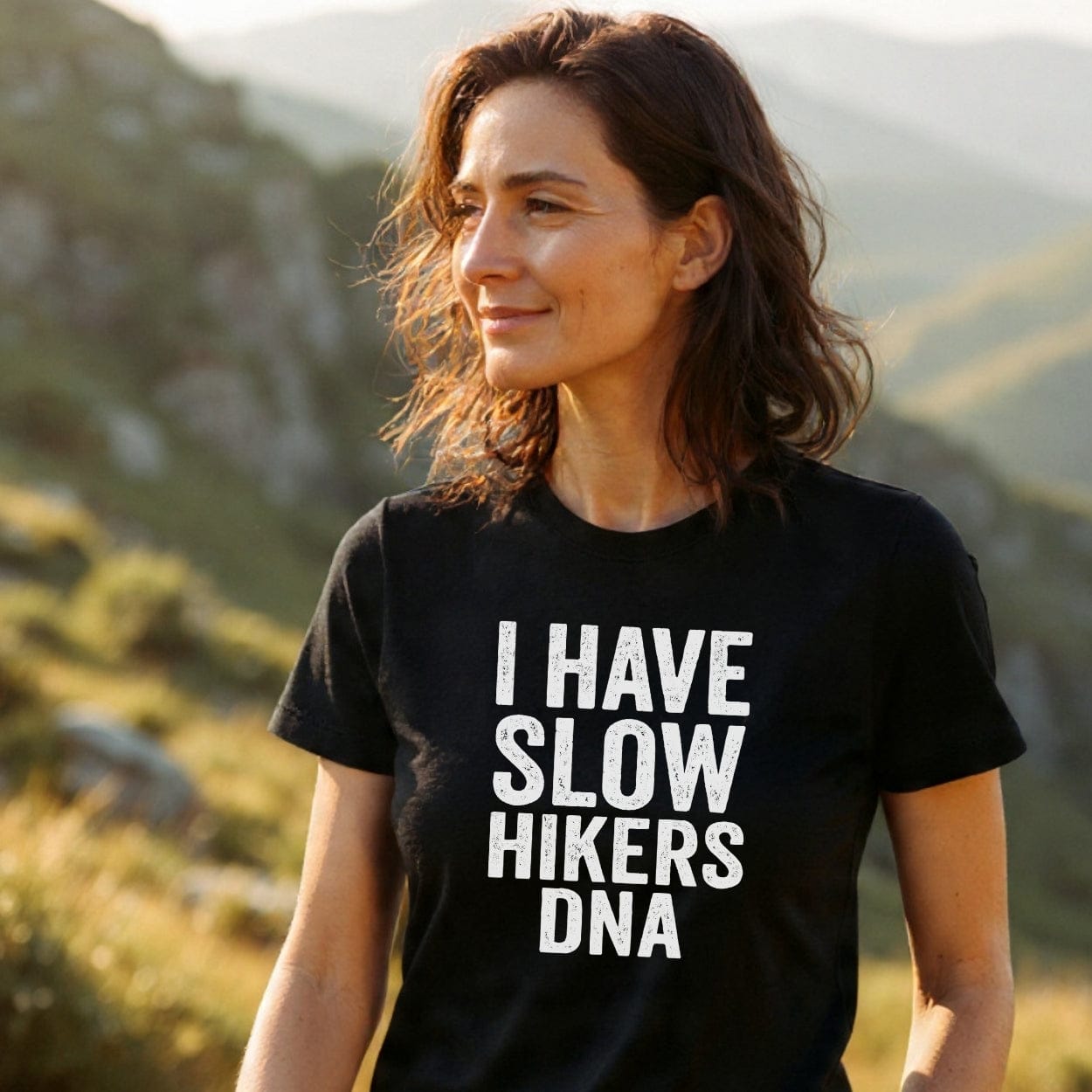 Woman outdoors in hilly landscape wearing a Slow Hikers DNA T-Shirt, featuring bold white text on black. She enjoys nature, exemplifying the shirt's blend of comfort, durability, and adventure spirit.