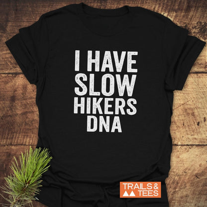 Slow Hikers DNA T-Shirt on wooden surface, featuring distressed white lettering and an orange “TRAILS & TEES” logo, styled with a pine sprig, highlighting outdoor adventure and comfort.