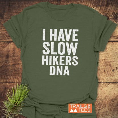 Olive-green T-shirt with bold white slogan “I HAVE SLOW HIKERS DNA” and orange “TRAILS & TEES” logo, crafted from soft ring-spun cotton, offering trail-tested comfort and adventure-ready design.