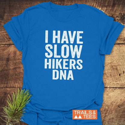 Slow Hikers DNA T-Shirt in bright blue with white distressed lettering, laid on wood. Features a small orange “TRAILS & TEES” label and an evergreen sprig accentuating its nature theme.