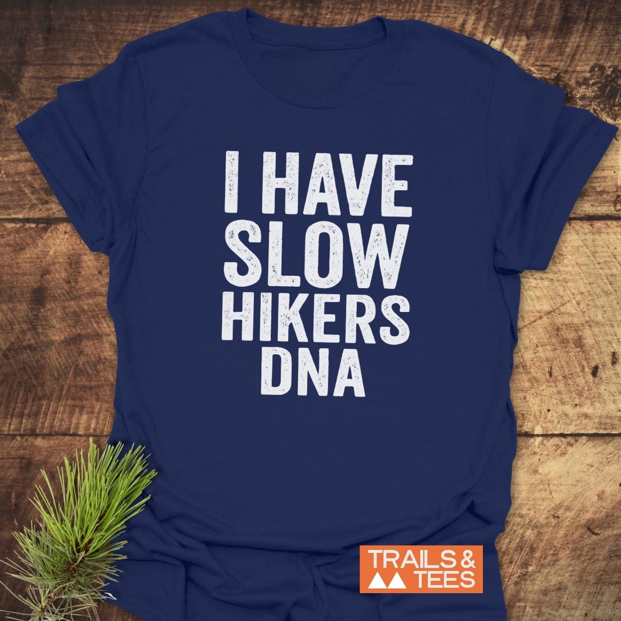 Slow Hikers DNA T-Shirt on wood surface, featuring a distressed white slogan. Includes a Trails & Tees logo and a pine sprig, highlighting its outdoor-inspired design and craftsmanship.