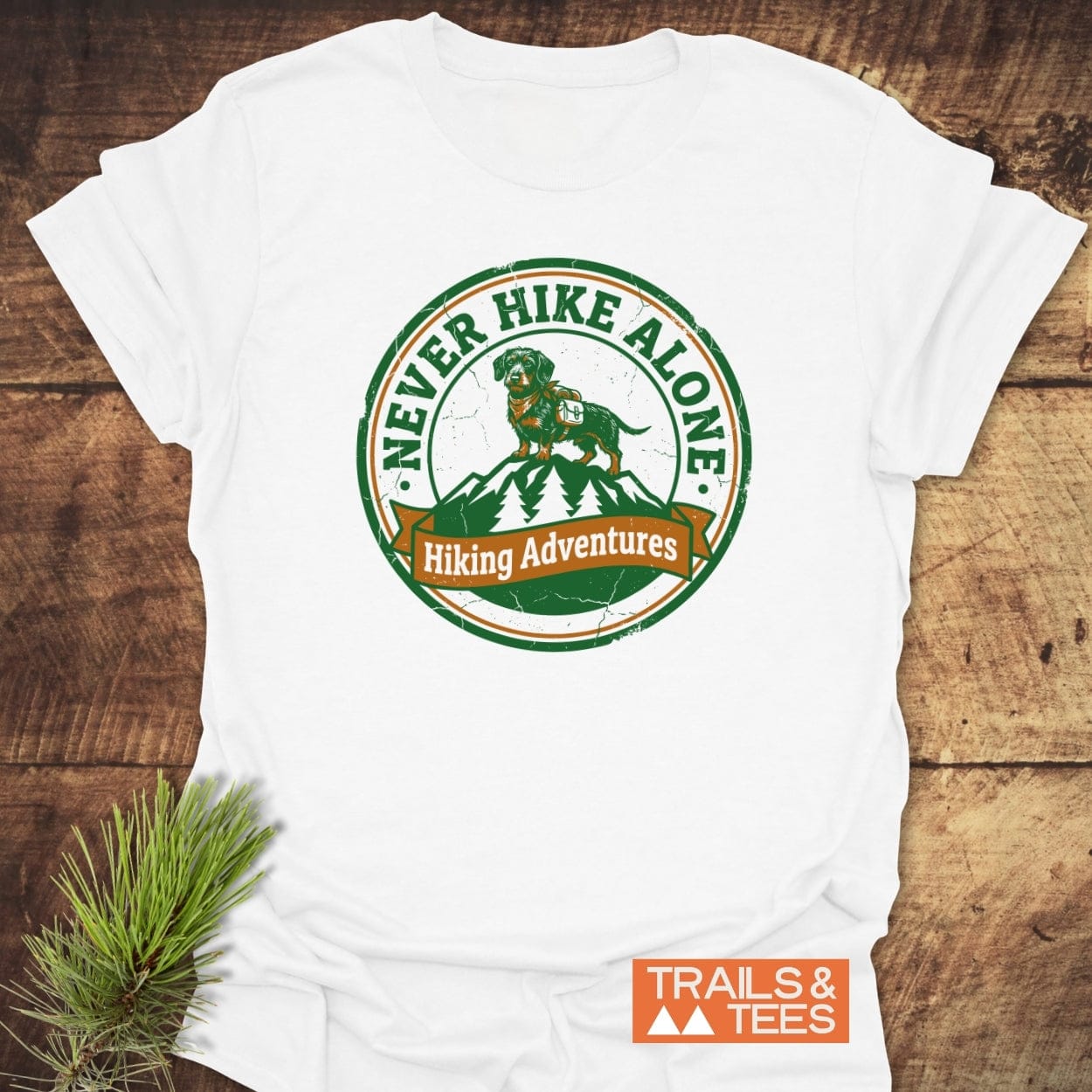 Never Hike Alone T-Shirt featuring a circular crest with a Dachshund on a mountain, surrounded by pine trees. Includes Hiking Adventures banner and TRAILS & TEES logo, laid on wood.