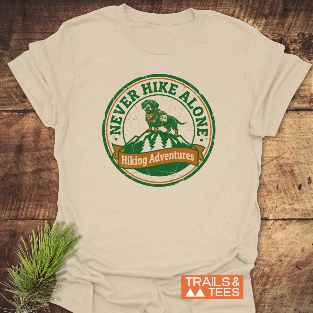 Never Hike Alone T-Shirt features a dachshund with a backpack on a mountain badge, symbolizing outdoor adventures. Crafted from lightweight cotton, it offers comfort and durability, perfect for nature enthusiasts.