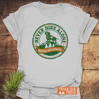 Never Hike Alone T-Shirt featuring a vintage-style badge with a dog on a mountain peak, laid on a wooden background. Includes Hiking Adventures banner and small TRAILS & TEES logo beside a pine branch.