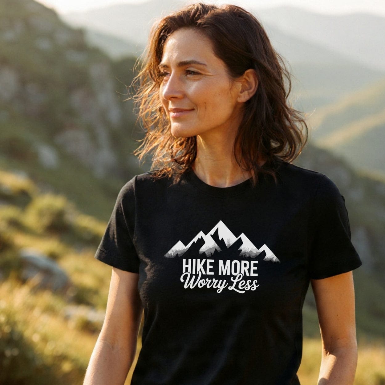 Woman wearing the Hike More Worry Less T-Shirt with a mountain graphic, set against a rolling hills backdrop, emphasizing adventure-ready comfort and durability for outdoor enthusiasts.