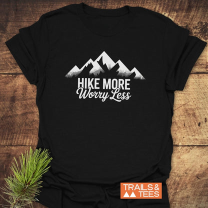 Black Hike More Worry Less T-Shirt with white mountain graphic, laid flat on wood. Features bold slogan, TRAILS & TEES logo, crafted from soft, lightweight cotton for outdoor adventures.