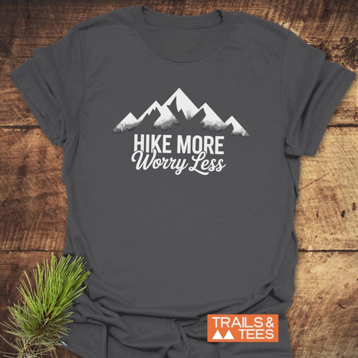 Charcoal-gray Hike More Worry Less T-shirt with mountain silhouette in white, displayed on wood with a pine sprig; embodies outdoor adventure spirit, crafted from soft, ring-spun cotton for comfort.
