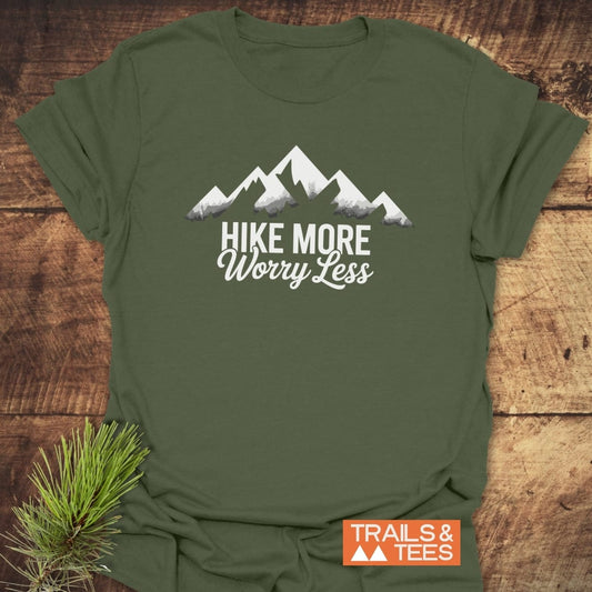 Olive-green Hike More Worry Less T-shirt with white mountain graphic, laid flat on wood. Includes small pine sprig and TRAILS & TEES logo in corner.