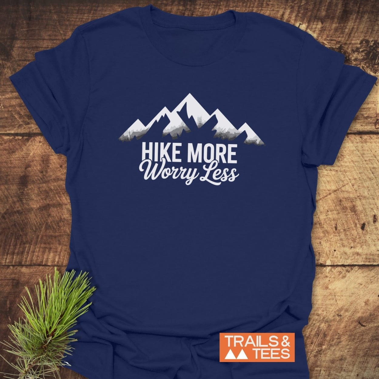 Navy-blue Hike More Worry Less T-shirt featuring a white mountain graphic, laid on wood. Includes Trails & Tees logo, and a pine sprig accent.