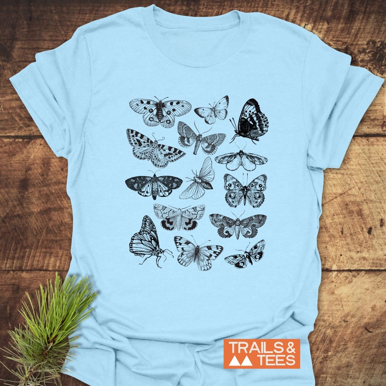 Vintage Butterfly Tattoo T-Shirt featuring intricate black line drawings of butterflies and moths on light-blue fabric, laid on a wooden surface with a TRAILS & TEES logo nearby.