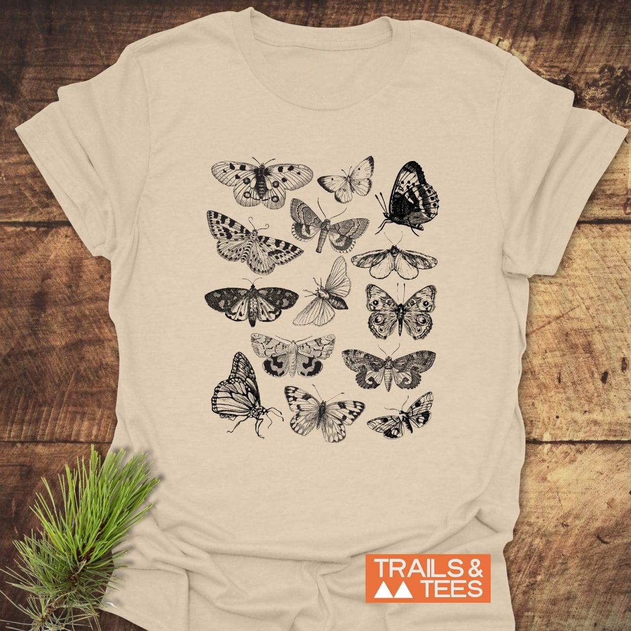 Vintage Butterfly Tattoo T-Shirt with intricate black butterfly and moth illustrations on beige fabric, perfect for nature enthusiasts. Laid on a rustic wooden background with a pine sprig beside it.