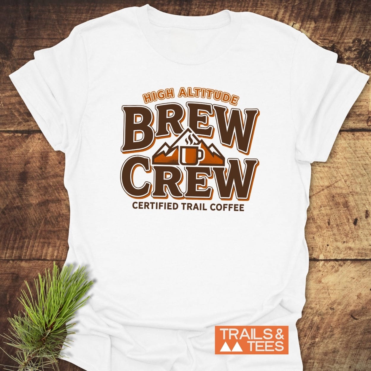 Trail Coffee T-Shirt featuring a mountain and coffee mug graphic with text, laid on wood with pine sprig, embodies outdoor adventure. Made from ring-spun cotton, it offers comfort and durability.