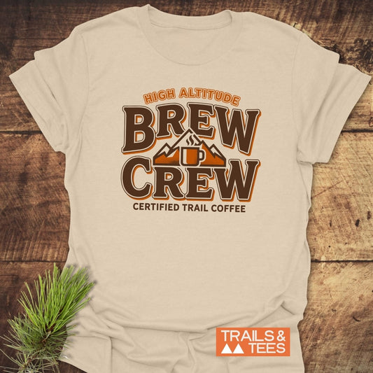 Trail Coffee T-Shirt featuring HIGH ALTITUDE BREW CREW logo with a coffee mug and mountain peaks, displayed flat on wood, embodies outdoor adventure spirit with durable, trail-inspired design.