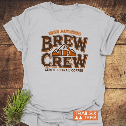 Trail Coffee T-Shirt featuring HIGH ALTITUDE BREW CREW with a coffee mug and mountains, displayed flat with pine sprig, illustrating trail-tested comfort and adventure-ready design from Trails & Tees.