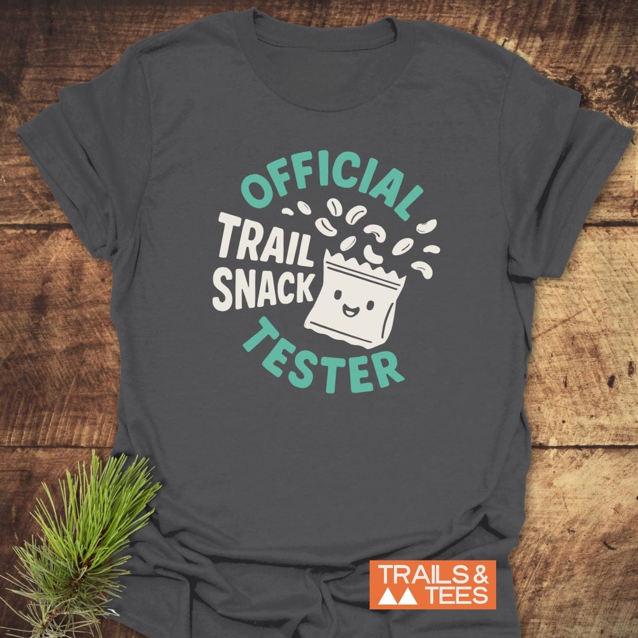 Official Trail Snack Tester T-Shirt with a smiling snack bag graphic and playful lettering, laid flat on wood. Features durable print, soft ring-spun cotton, and designed for adventurous outdoor enthusiasts.